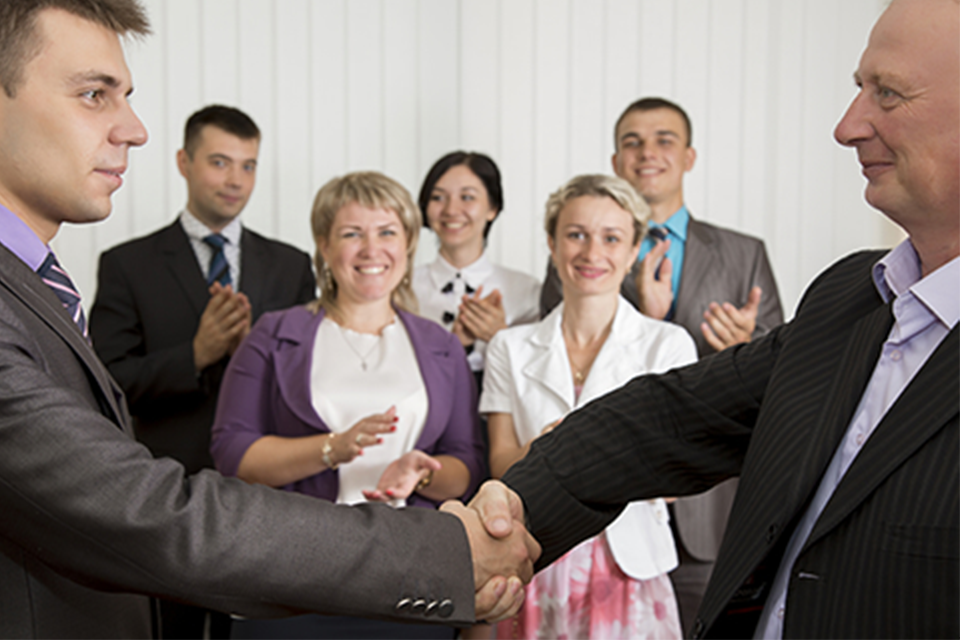 Image of two men shaking hands in the foreground with a small group clapping in the background.